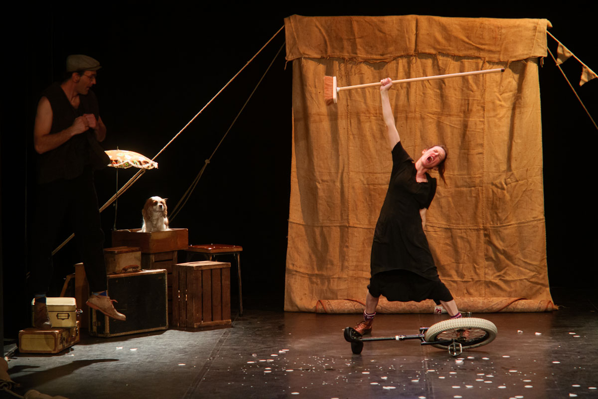Sur une scene de theatre. Au centre, une artiste en robe noire des annees 30 poing levé avec un balai dans la main. Coté jardin, un artiste bondit. Au centre de la piste, un monocycle posé au sol. En fond, un chien sur des valise regarde la scene.